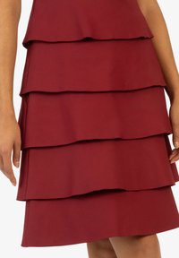 Burgunderfarbenes, geschichtetes Kleid aus weichem, glattem Stoff mit vier Rüschenschichten und einem taillierten Oberteil. Eine Nahaufnahme des Designs betont die Textur und Form.