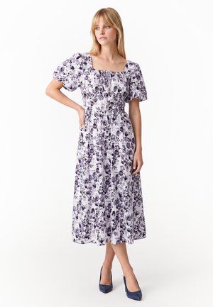 Donna bionda che indossa un vestito midi bianco con stampa floreale viola, maniche a sbuffo, scollo quadrato e tacchi blu navy, in piedi con una mano sul fianco.
