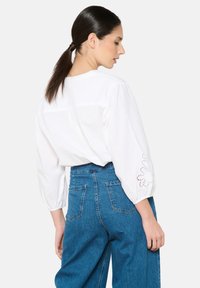 Blusa de algodón blanca con mangas globo de 3/4 de largo que presenta detalles florales bordados. Combinada con jeans de mezclilla azul de tiro alto.