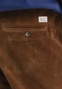 Brown corduroy pants featuring a back pocket with a button closure and a tag displaying the brand "GANT." Accented with textured ribbing.