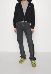 Osoba v černé bundě, šedém tričku, tmavě šedých džínách, neonově žlutých a hnědých teniskách a pásku s neonově zeleným textem.