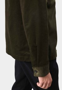 Oliwkowa dżinsowa kurtka o prostym kroju, z detalami przy mankietach na guziki i teksturowanym wykończeniem, noszona na ciemnych spodniach.