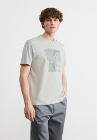 Νέος άνδρας με κυματιστά μαλλιά, φορούν ανοιχτό γκρι μπλουζάκι με στάμπα και γκρι παντελόνι, στέκεται μπροστά σε απλό λευκό φόντο.