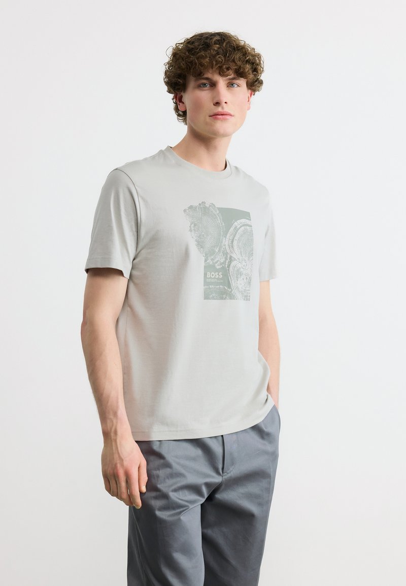 Νέος άνδρας με κυματιστά μαλλιά, φορούν ανοιχτό γκρι μπλουζάκι με στάμπα και γκρι παντελόνι, στέκεται μπροστά σε απλό λευκό φόντο.