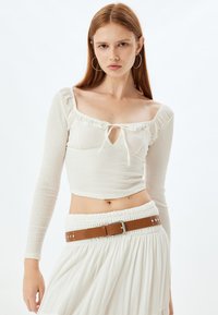 Witte lange mouw crop top met een vierkante halslijn, geplooide details en een strik in het midden. Te combineren met een witte rok en een bruine riem. Gladde textuur.