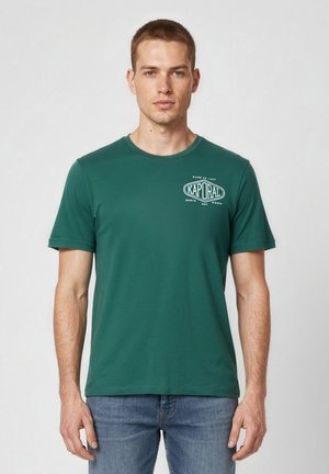 Man draagt een groen Kaporal T-shirt met korte mouwen en blauwe jeans, staand tegen een effen lichte achtergrond.