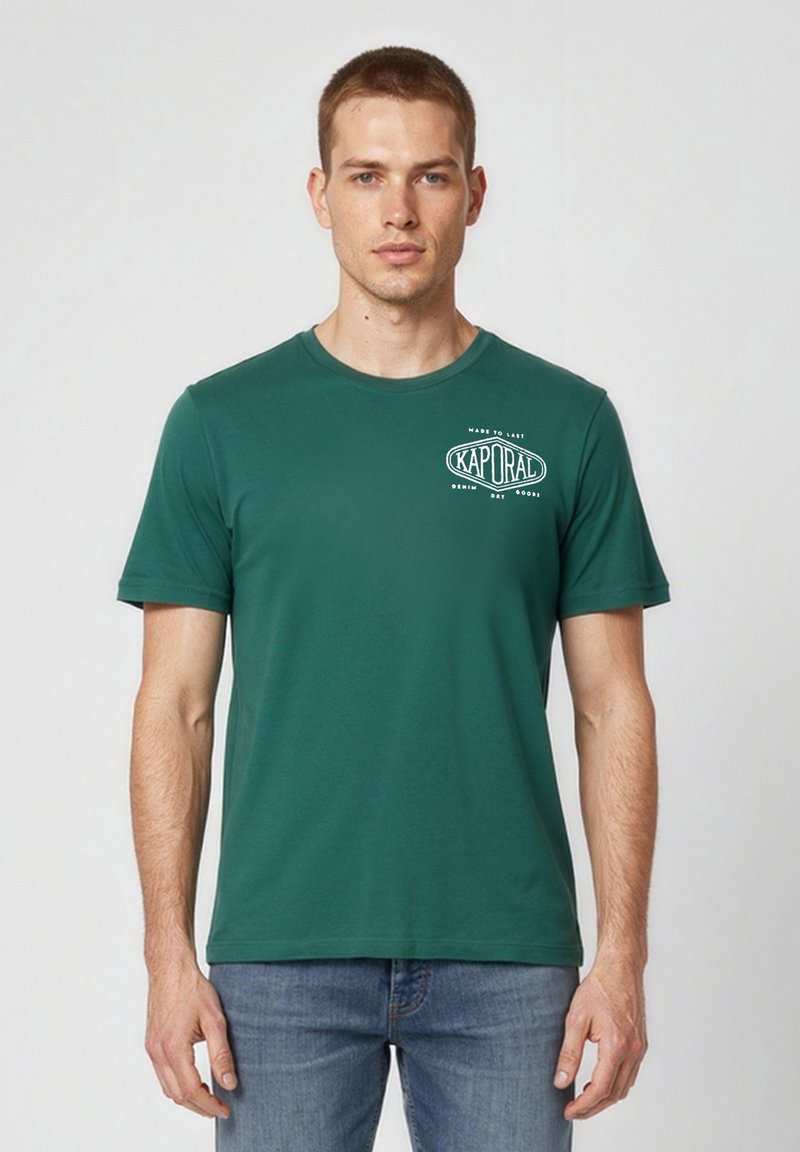 Homme portant un t-shirt vert à manches courtes de la marque Kaporal et un jean bleu, debout devant un fond uni clair.