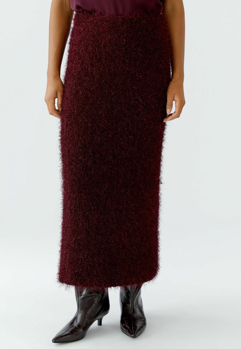 Femme portant une longue jupe en peluche rouge foncé associée à des bottines noires brillantes à bout pointu, se tenant contre un fond uni.