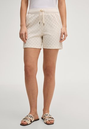 Donna che indossa pantaloncini crema con motivo e coulisse e sandali beige a ciabatta con scritta nera "JOOP!", in piedi contro uno sfondo neutro.