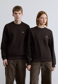 Καφέ φούτερ με ριμπ μανσέτες και τελειώματα, με ένα μικρό πράσινο λογότυπο στο αριστερό στήθος. Τα μοντέλα φορούν ταιριαστά καφέ παντελόνια.