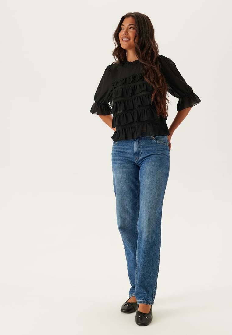 Blusa nera con ruches e maniche lunghe a campana, abbinata a jeans blu a gamba dritta e ballerine nere, in piedi su uno sfondo neutro.