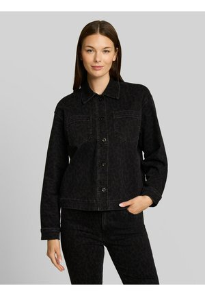 Vrouw met lang bruin haar draagt een zwarte jas met knopen en bijpassende broek met subtiel luipaardprint, staand tegen een effen achtergrond.