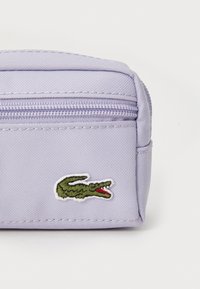 Pochette en tissu violet clair avec fermeture à glissière et logo de crocodile vert. Texture lisse, design compact, adaptée pour de petits objets.