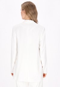 Bílý blazer s dlouhými rukávy a přiléhavým střihem. Má hladkou texturu a jednoduchý design, který sahá k polovině stehna vzadu.