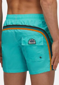 Shorts de bain turquoise en tissu léger et à séchage rapide. Caractéristiques : rayures noires et orange, une poche au dos et un patch logo SUNDEK.