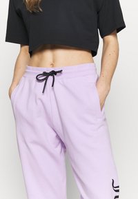 Fialové tepláky s elastickým pasem a černou šňůrkou; mají boční kapsy a černé logo na levé noze.