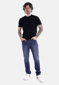 T-shirt nera a maniche corte, jeans in denim blu, sneakers bianche con dettagli neri. Il modello ha dei tatuaggi e sta in piedi con le mani in tasca.