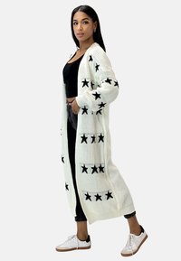 Cardigan lavorato a maglia color crema con motivi neri a forma di stella, apertura frontale, maniche lunghe e polsini a costine, abbinato a una maglietta nera, jeans e sneakers bianche.