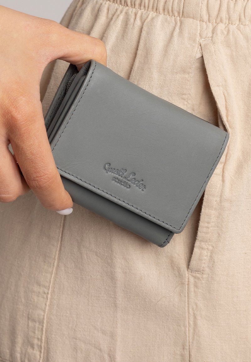 Pilkos odos piniginė su lankstymo dizainu, su matomomis siūlėmis ir įspaustu logotipu, laikoma prieš šviesiai rudos audinio foną.