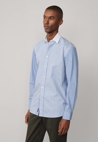 Camisa de botón azul claro con rayas blancas verticales, cuello clásico, mangas largas y una etiqueta en el dobladillo. Textura de tela suave.