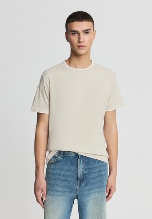 Maglietta beige con scollo rotondo e maniche corte, caratterizzata da un colletto bianco a contrasto. Indossata con jeans di denim azzurro chiaro, che mostrano una texture scolorita.