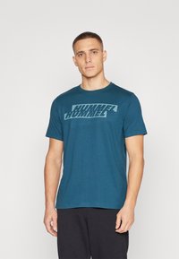 Türkise Baumwoll-T-Shirt mit kurzen Ärmeln, das vorne in heller Blaufarbe mit einem großen, grafischen "HUMMEL"-Druck in der Mitte versehen ist. Klassisches Rundhalsdesign.