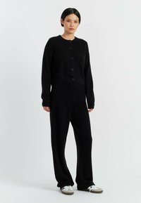 Zwarte gebreide cardigan met ronde hals en knoopsluiting, gecombineerd met bijpassende zwarte brede broek. Het model draagt witte sneakers.