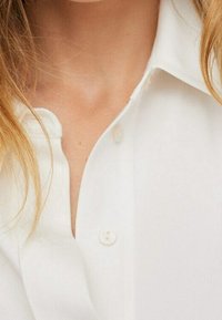 Weiße Bluse mit Kragen, ausgestattet mit einer klassischen Knopfleiste und einer glatten Stoffstruktur. Nahaufnahme, die den Ausschnitt hervorhebt.