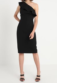 Schwarzes, ein Schulter-Kleid mit gerafftem Ausschnitt. Figurbetonte Silhouette, kniebedeckt, kombiniert mit schwarzen Riemchen-High Heels. Glatte Stofftextur.