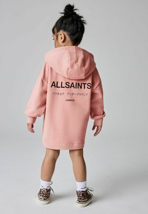 Sweat-shirt à capuche rose avec logo sur le devant et design au dos. Porté avec des baskets à motifs dotées d'accents noirs et marron.