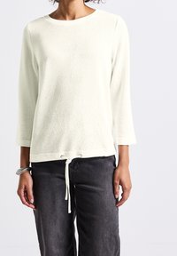 Haut à manches longues texturé couleur crème claire avec un col rond et un ourlet à cordon de serrage. Associé à un jean gris ample.