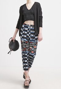 Schwarzer strukturierter Cardigan über einem schwarzen gemusterten Crop-Top, kombiniert mit mehrfarbigen gestreiften Hosen. Modell hält eine runde schwarze Tasche. Schwarze High Heels.