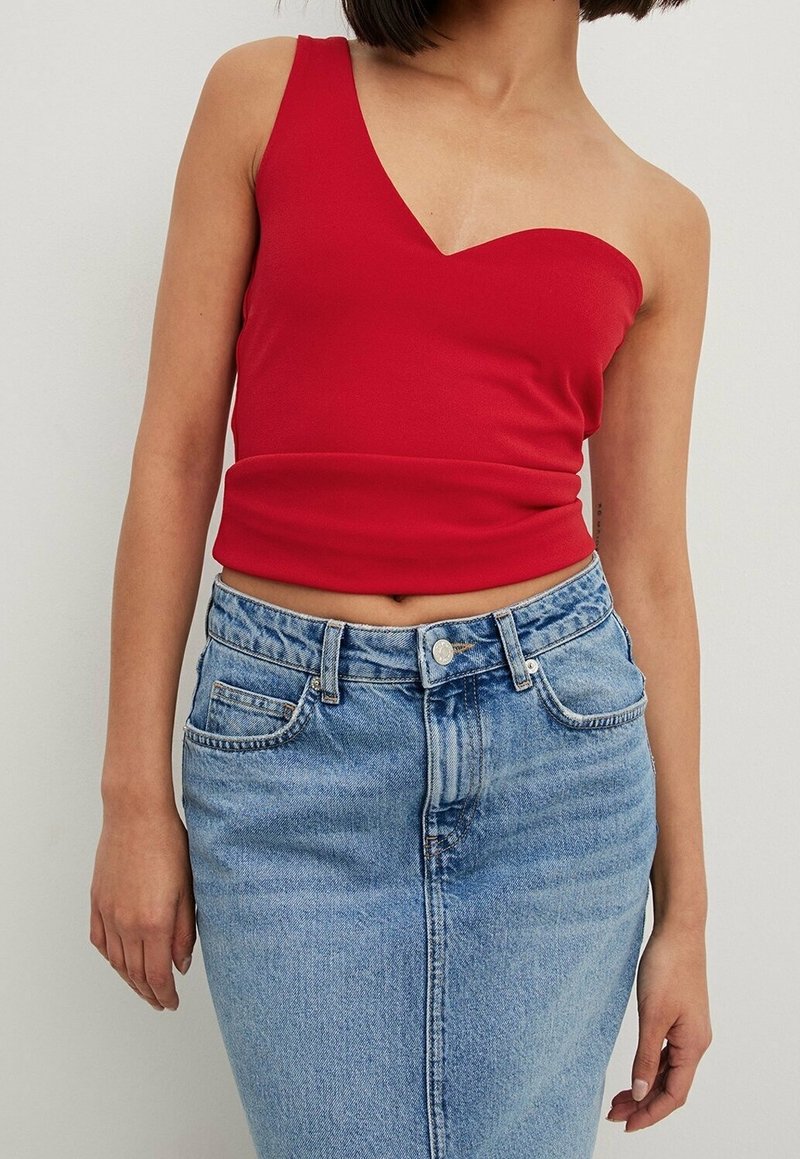 Femme portant un crop top rouge à une épaule et une jupe en jean bleue taille haute, debout devant un fond clair uni.