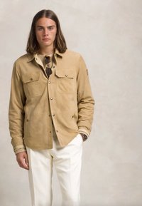 Καφέ σουέτ μπουφάν με δύο τσέπες στο στήθος και κουμπιά με κούμπωμα, συνδυασμένο με κρεμ ζακέτα από πλέγμα και λευκά παντελονιά. Ορατό διακοσμητικό ρολόι.