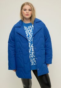 Giacca imbottita blu con un ampio colletto, tasche frontali e chiusura con bottoni. Indossata sopra un maglione blu a fantasia e leggings neri.