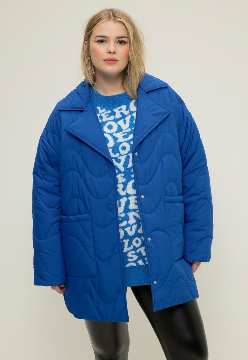 Giacca imbottita blu con un ampio colletto, tasche frontali e chiusura con bottoni. Indossata sopra un maglione blu a fantasia e leggings neri.