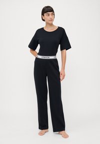 Zwart katoenen jumpsuit met een ronde halslijn, korte mouwen en een witte, gebrandmerkte elastieken tailleband. De broekspijpen zijn wijd en losvallend.