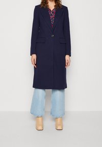 Marineblå uldfrakke med skræddersyet design, enkelt knap, hakrevers og to forlommer, kombineret med lyse denimbukser med vidde og beige hæle.