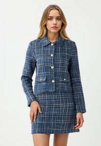 Młoda kobieta w niebiesko-białą kratkę, nosząca zapinaną na guziki kurtkę z złotymi guzikami oraz pasującą spódnicę, stoi na tle jednolitego tła.