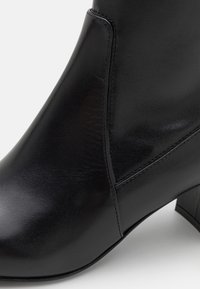 Gros plan sur une bottine noire en cuir lisse avec des coutures apparentes et un talon bloc sur fond clair.