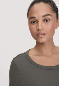 Junge Frau mit glatter Haut und zurückgegebene Haare, die ein olivgrünes, geripptes Oberteil mit einem runden Ausschnitt trägt, vor einem einfarbigen hellen Hintergrund.