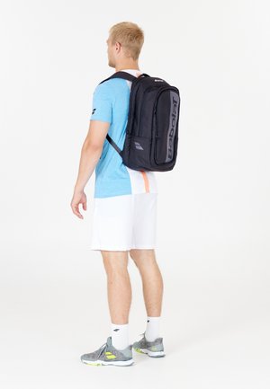Zaino Babolat nero con logo, forma rettangolare, spallacci imbottiti e molteplici scomparti. Indossato da una persona in camicia blu e pantaloni bianchi.