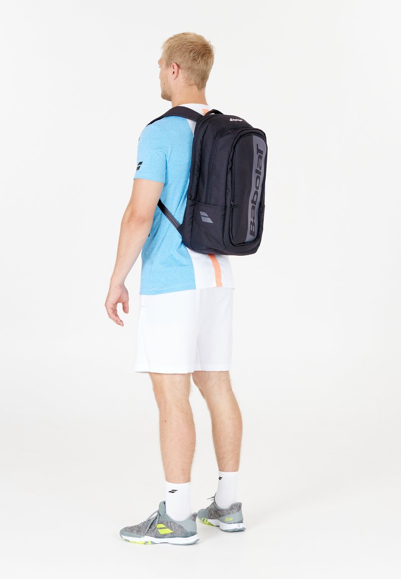 Mochila negra de Babolat con logo, forma rectangular, correas acolchadas para los hombros y múltiples compartimentos. Llevada por una persona con una camisa azul y pantalones cortos blancos.