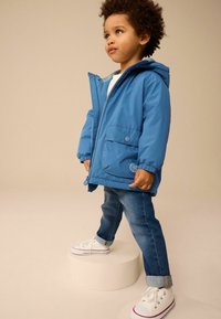 Blauwe waterdichte jas met een capuchon, voorrits en drukknoopzakken, gecombineerd met spijkerbroeken en witte sneakers. Eenvoudig en functioneel ontwerp.