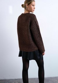 Jeune femme aux cheveux blonds attachés en chignon, portant un pull marron texturé, une jupe noire et des collants noirs, debout devant un mur clair uni.