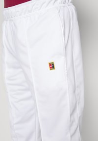 Pantalones deportivos blancos con una cintura elástica, que presentan franjas laterales y un pequeño parche de logo multicolor en el muslo izquierdo.