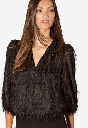 Vrouw met lang bruin haar, die een doorschijnende zwarte blouse draagt met textuurige verticale fringes en driekwart mouwen, kijkt opzij.