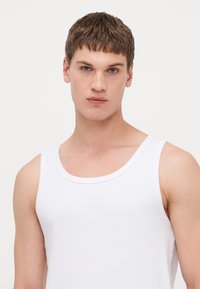 Valge puuvillane tank top ümmarguse kaelusega ja laia õlavarrega, millel on ribakujuline tekstuur ja keha järgi istuv disain.