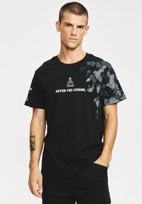 Czarna koszulka z krótkim rękawem z graficznym wzorem na jednym ramieniu oraz białym napisem „ENTER THE LEGEND.”, wykonana z bawełnianej tkaniny.