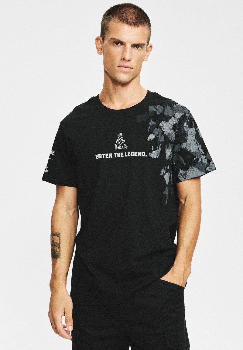 Czarna koszulka z krótkim rękawem z graficznym wzorem na jednym ramieniu oraz białym napisem „ENTER THE LEGEND.”, wykonana z bawełnianej tkaniny.